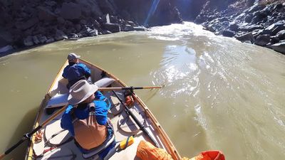 Overhead view from a wooden dory drifting through swirling boils in a calm eddy of Westwater Canyon's dark inner gorge, one boater glancing back as sunlight streaks off the water.