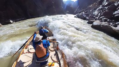 Wooden dory drops toward a stacked wave train in Sock-It-To-Me rapid, Westwater Canyon, dark boulders and sheer metamorphic walls framing the whitewater ahead.