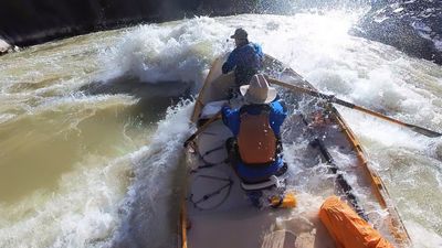 Wooden dory buries into breaking waves in the heart of Sock-It-To-Me rapid, whitewater exploding around both boaters between dark gorge walls.