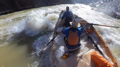 Wooden dory pitches through a violent section of Sock-It-To-Me rapid, an oar flung wide and whitewater breaking over the crew in Westwater Canyon's dark gorge.