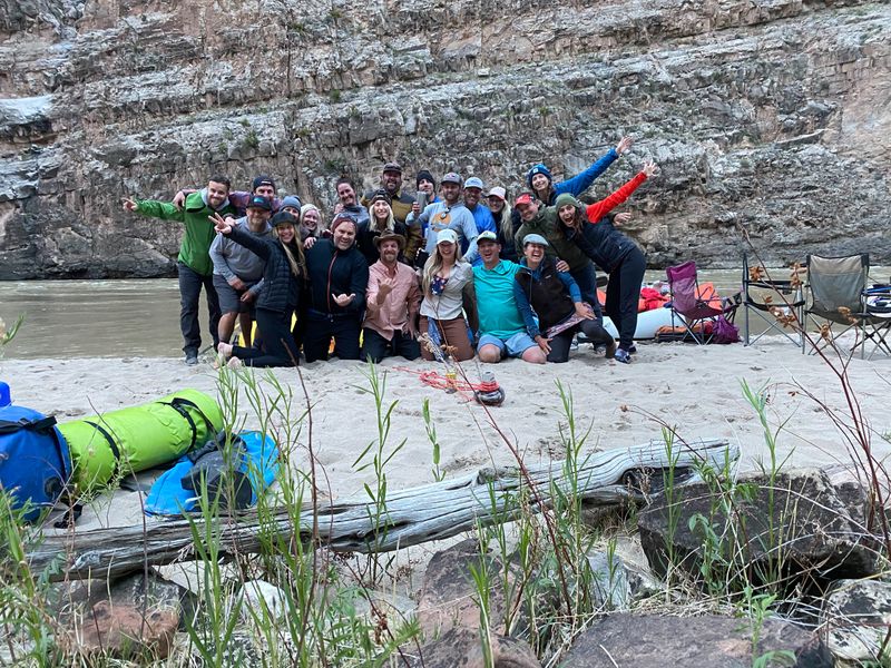 A large rafting group of roughly two dozen people crowds together for a portrait on a sandy beach below a fractured limestone canyon wall